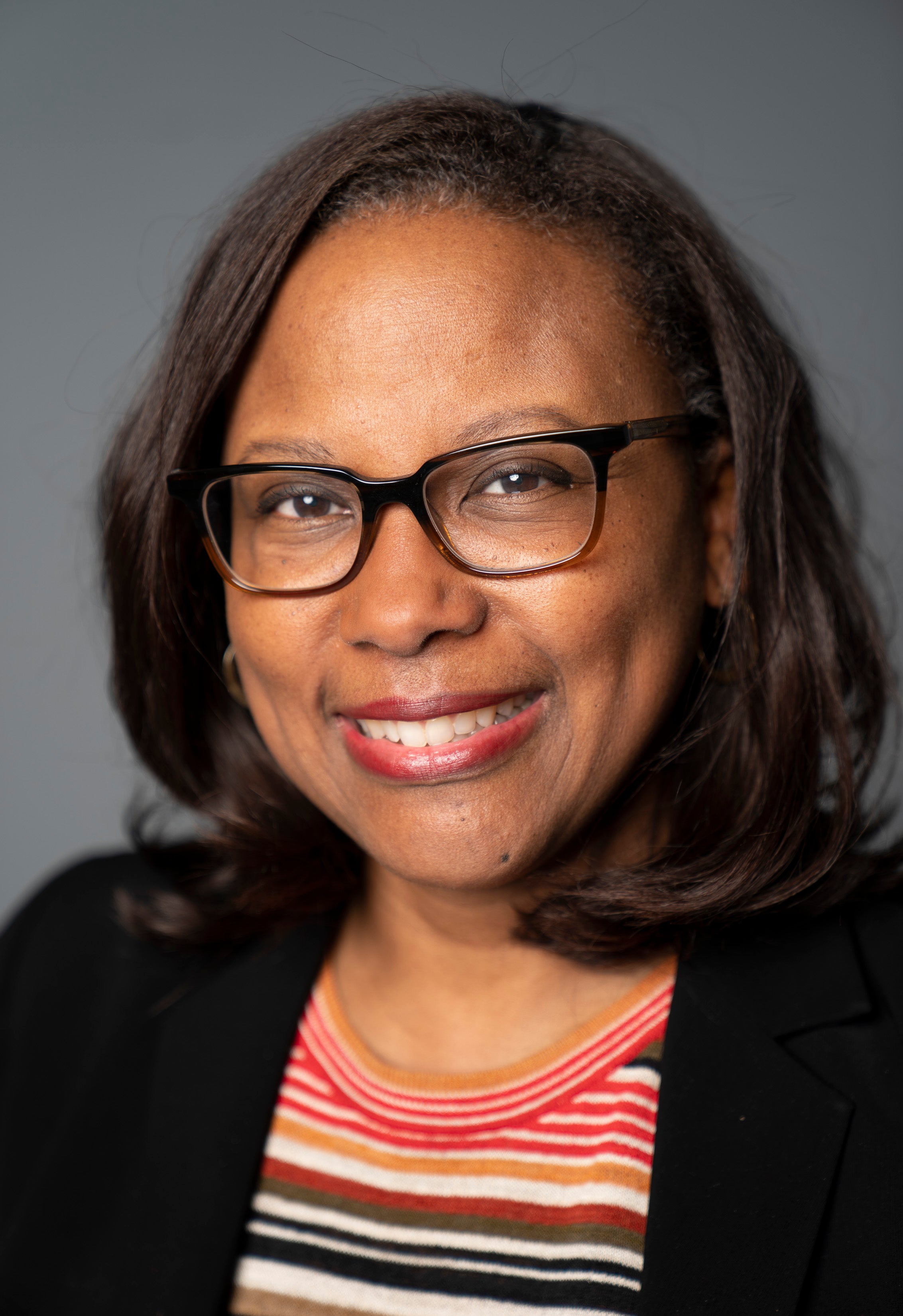 Portrait of Maria Reeve smiling warmly at the camera, wearing dark-framed glasses, a black blazer, and a striped top in red, orange, and white tones, against a neutral gray background.