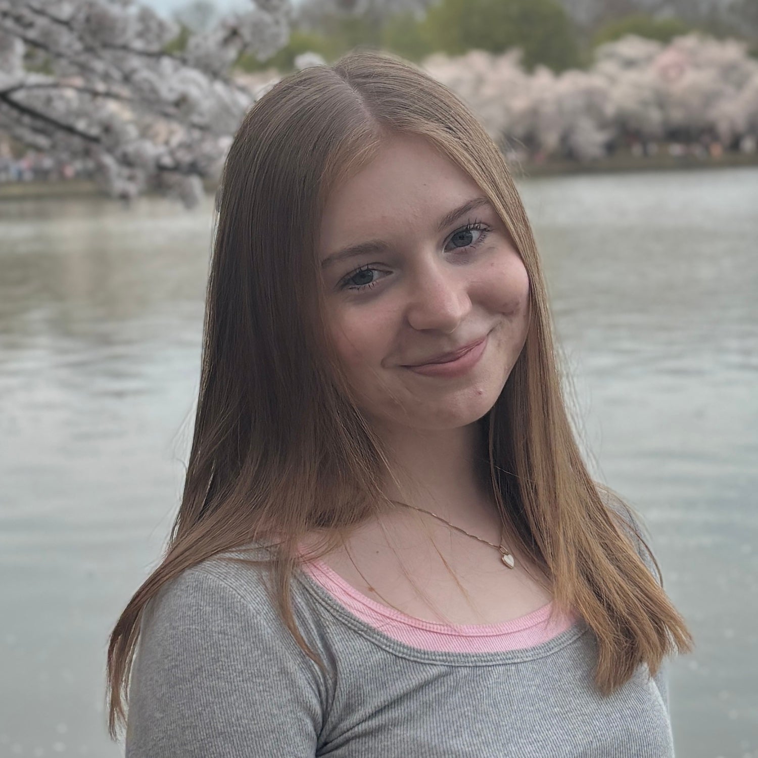 Portrait of Amelia Twyman standing by a calm body of water, wearing a gray top with a pink neckline and a delicate necklace, with blooming cherry trees softly blurred in the background.