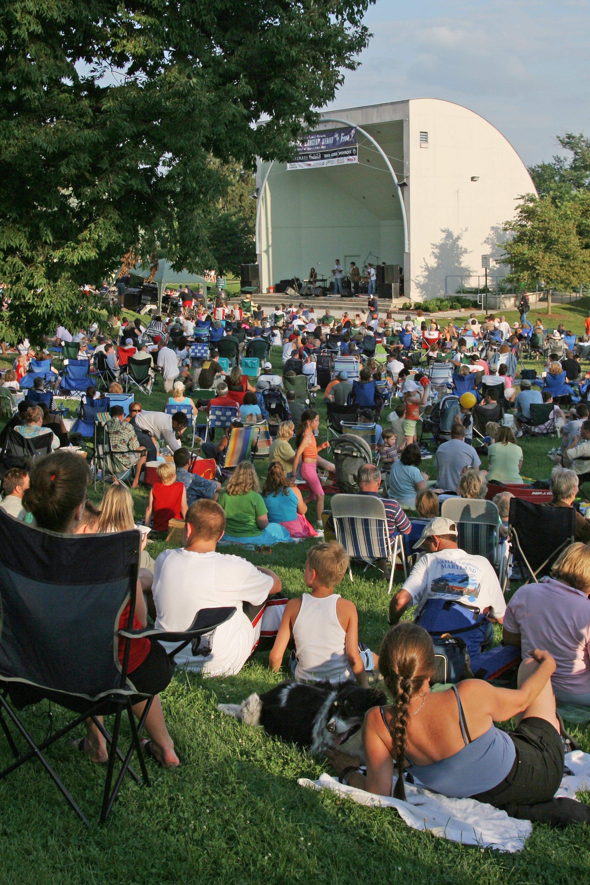 A crowd of people sitting on the grass listening to music at the Baker Park Bandshell in Frederick, Maryland.