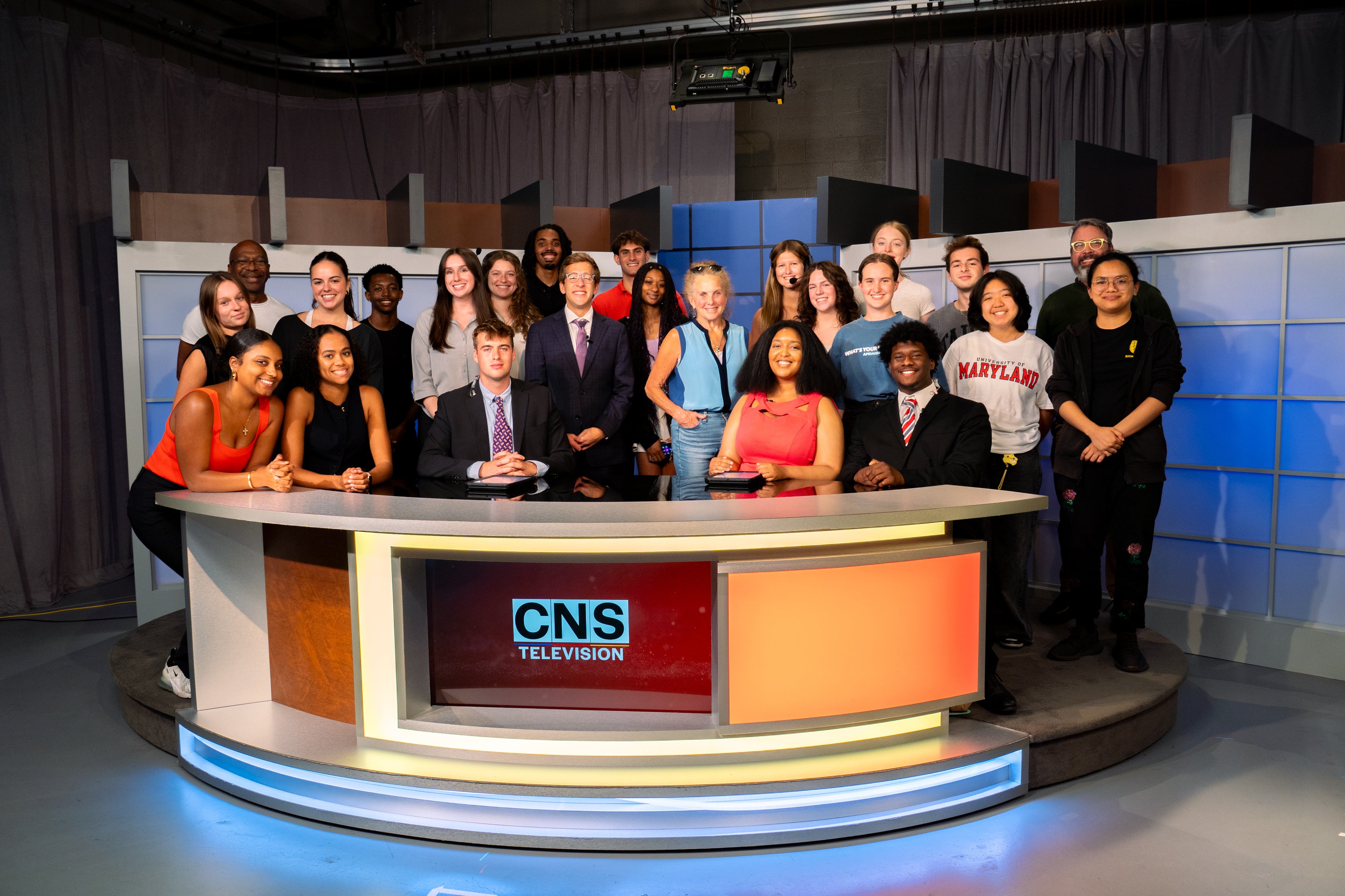 A large group poses on the CNS Television studio set behind and around a curved anchor desk with the CNS Television logo, under studio lights and blue-and-gray backdrop panels.