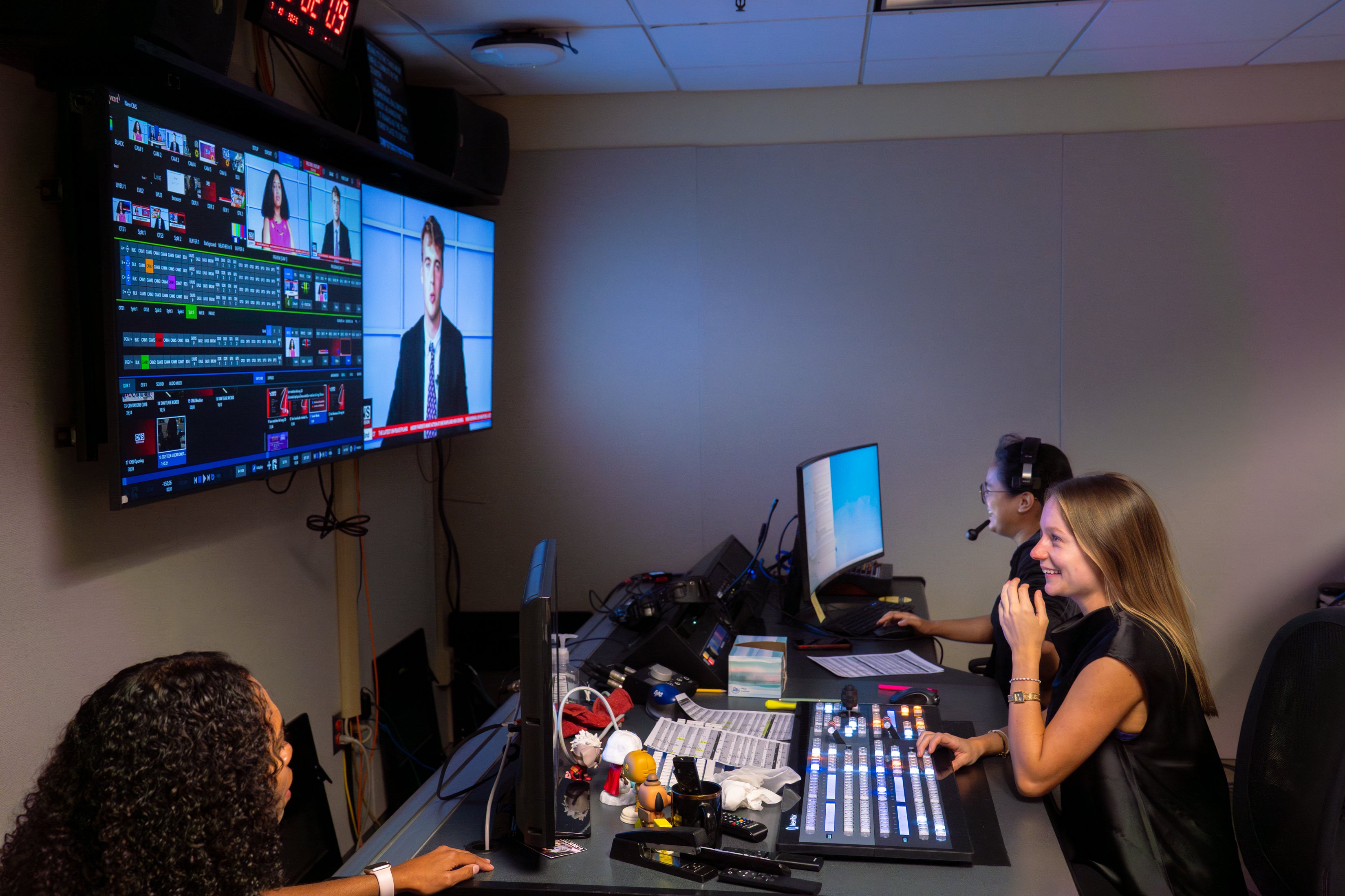 In a broadcast control room, three people work at computers and a video switcher while large monitors display a news anchor and production feeds.