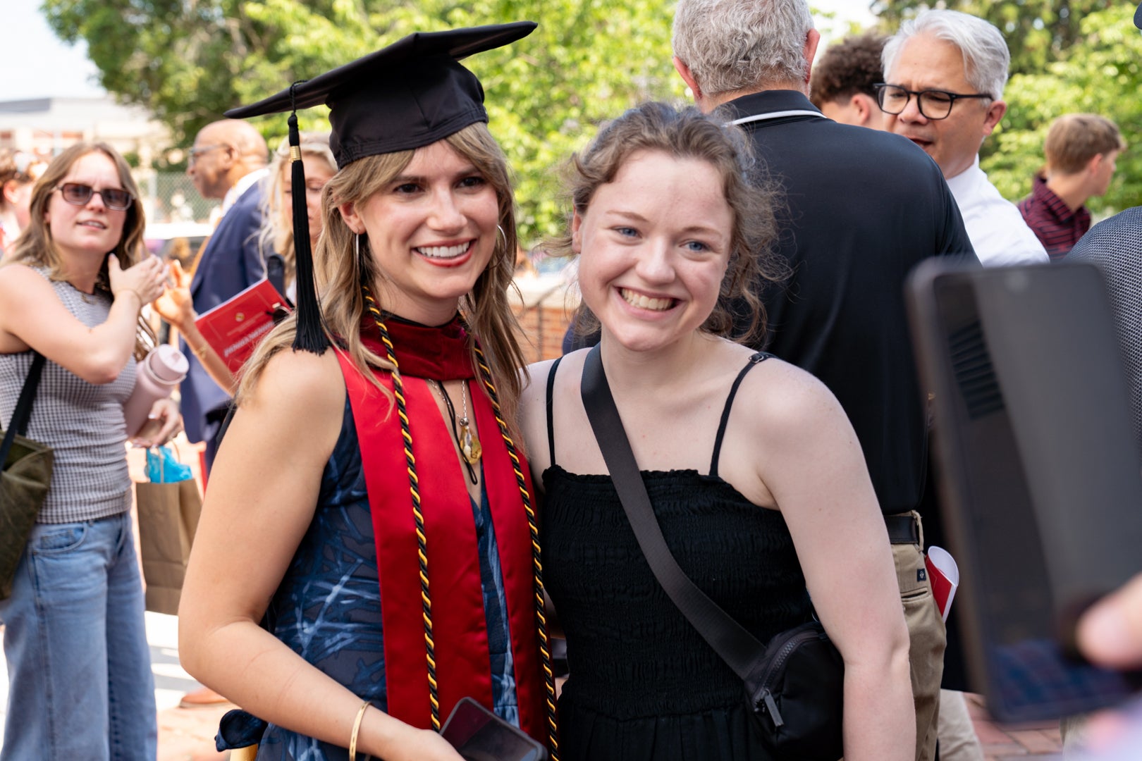 Outdoors after commencement, a graduate wearing a mortarboard, red stole, and honor cords stands arm-in-arm with a friend while people mingle behind them.