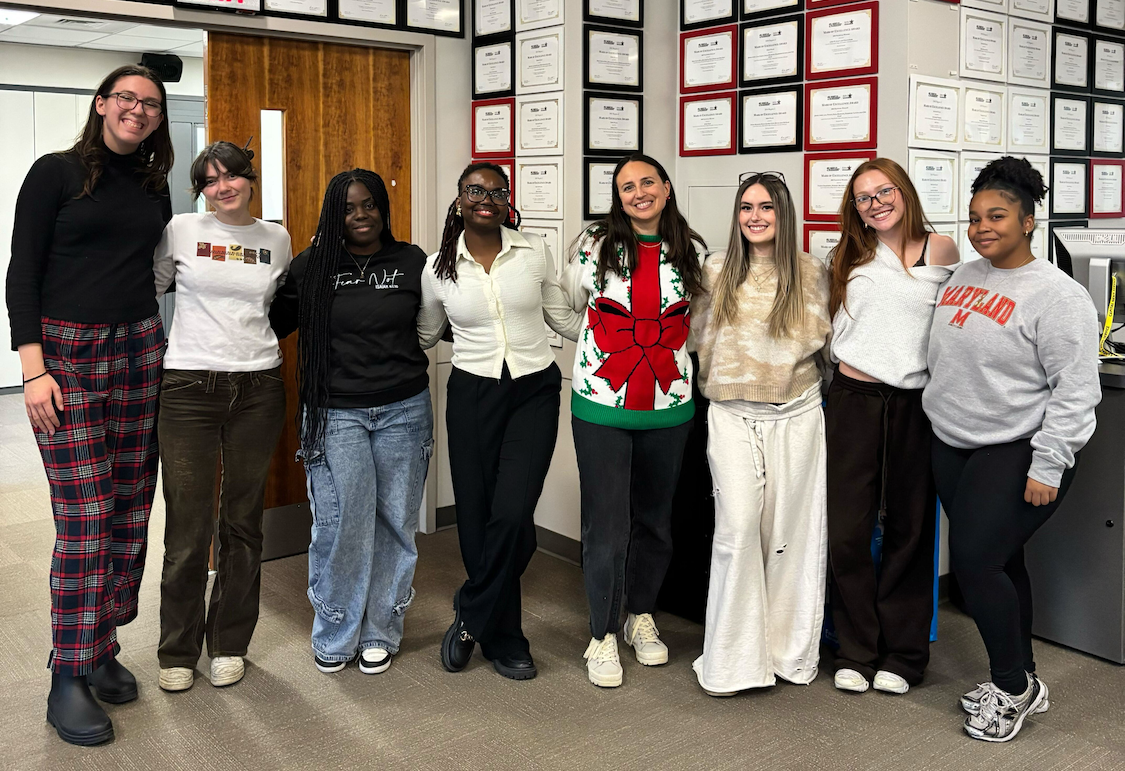 Members of the Capital News Service audience engagement bureau pose together in an office lined with framed awards and certificates. The group of eight students stands arm in arm, smiling at the camera, wearing a mix of casual and festive clothing, including a holiday sweater and University of Maryland apparel.