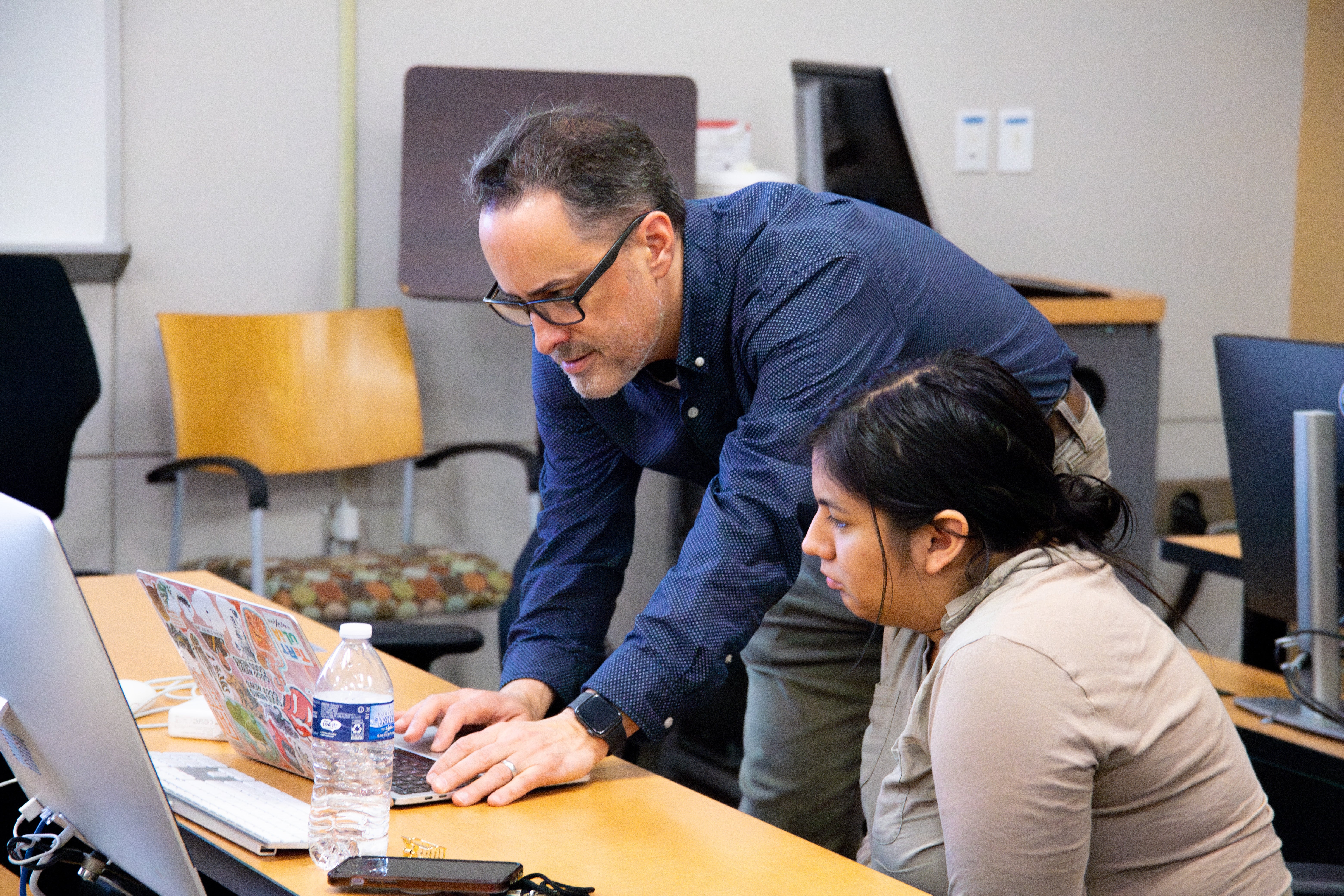 A man faculty member leans over a laptop to assist a woman student working at a computer in a classroom or newsroom workspace. Another monitor and laptop are visible on the table.