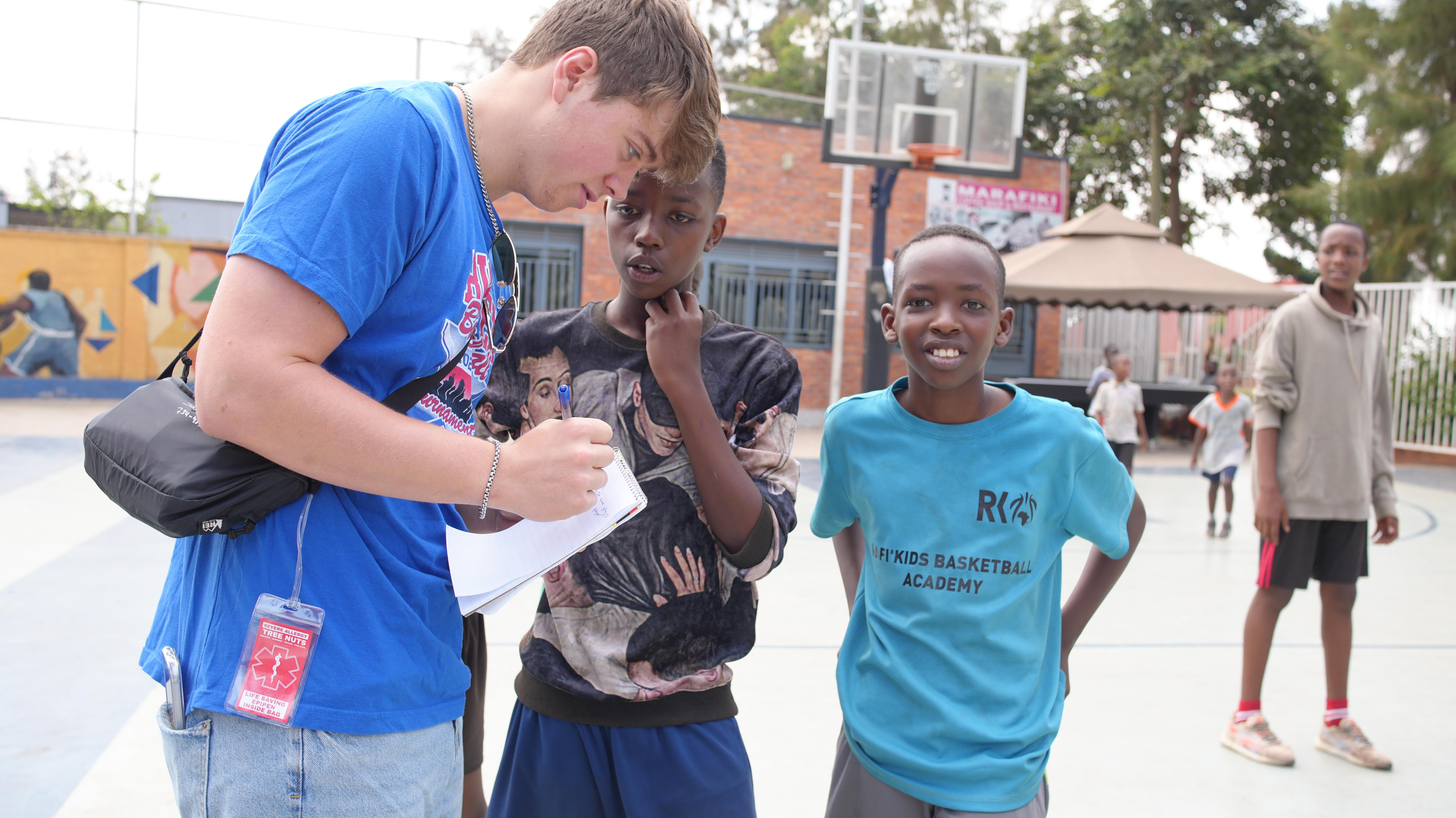 A student speaks with two boys on an outdoor basketball court. The student holds a notebook and pen, while one boy wears a “MFI Kids Basketball Academy” shirt. Other children and a basketball hoop are visible in the background.