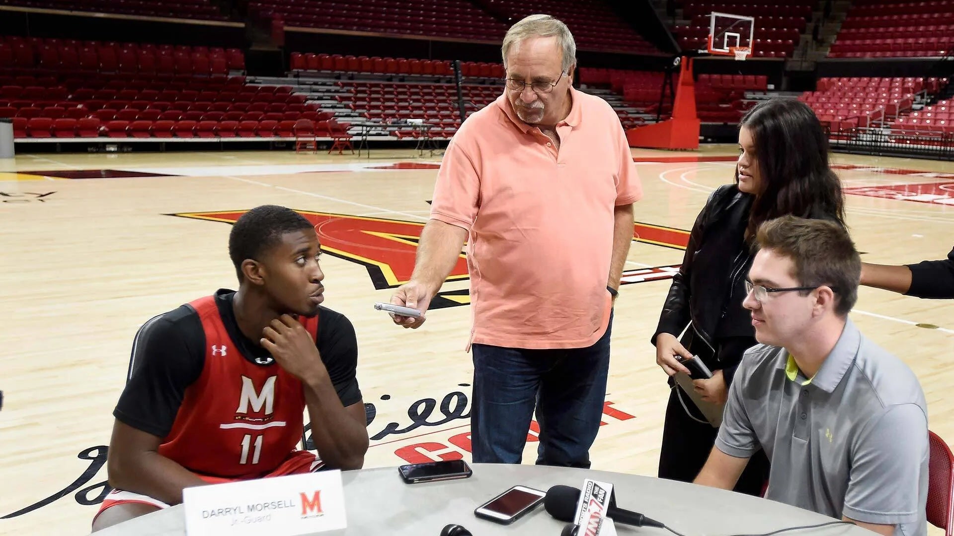 David Ginsburg (center), a 1975 alumnus of the University of Maryland’s Philip Merrill College of Journalism, interviews Maryland men’s basketball player Darryl Morsell on the court at Xfinity Center while two student journalists observe during a media session.