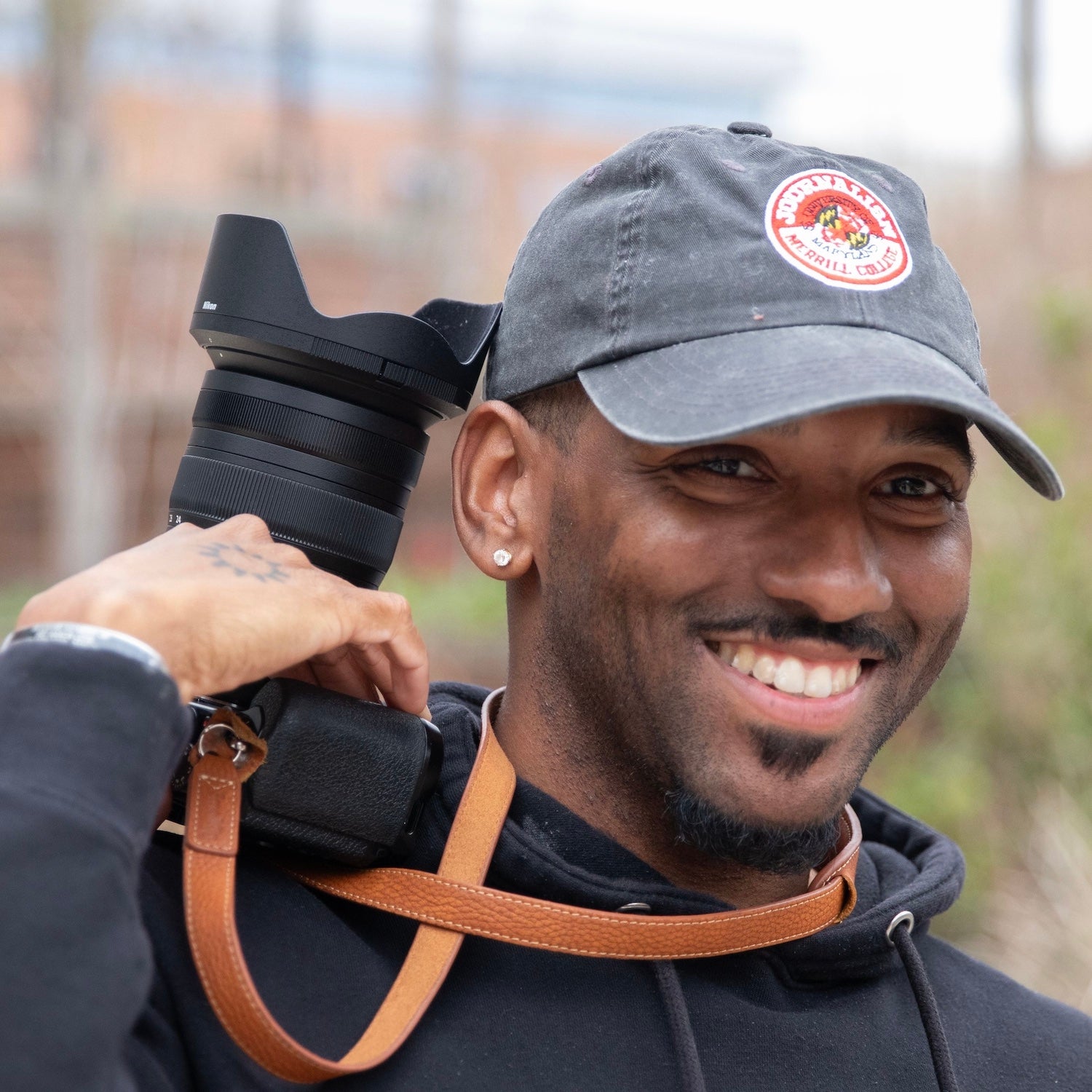 Portrait of Ryan Bowie outdoors, holding a camera with a large lens over his shoulder, wearing a dark hoodie and a gray baseball cap with a patch; the background is softly blurred.