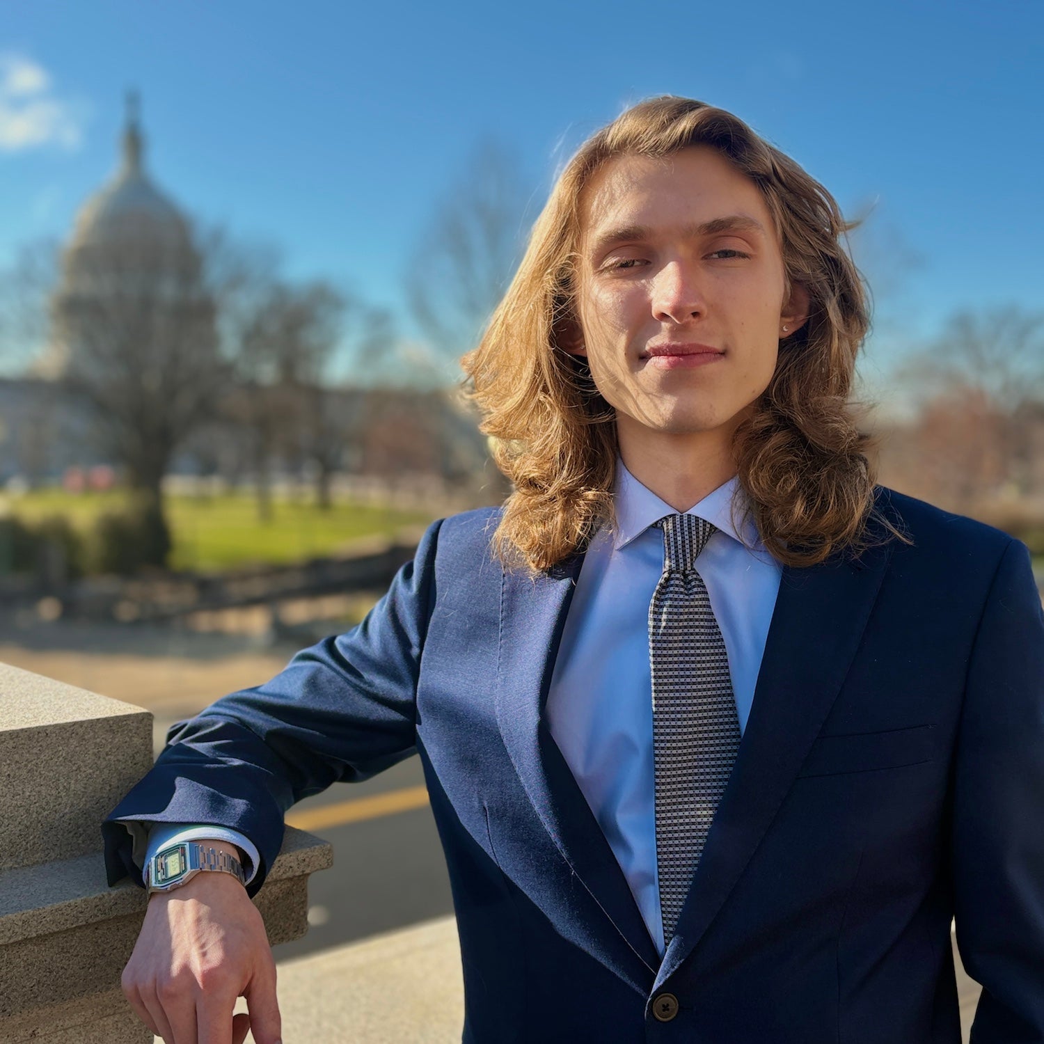 Portrait of Stephen Lotz outdoors on a sunny day, leaning on a stone railing in a navy suit, light blue shirt, and patterned tie, with the U.S. Capitol dome blurred in the background.