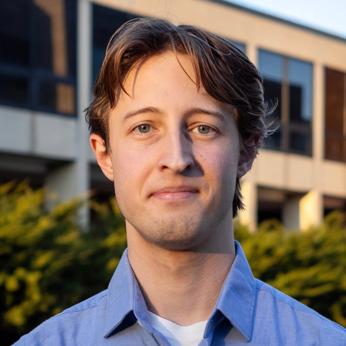 Portrait of Tyler Quattrin outdoors in front of a campus building, wearing a light blue collared shirt, with warm late-day sunlight and greenery softly blurred in the background.