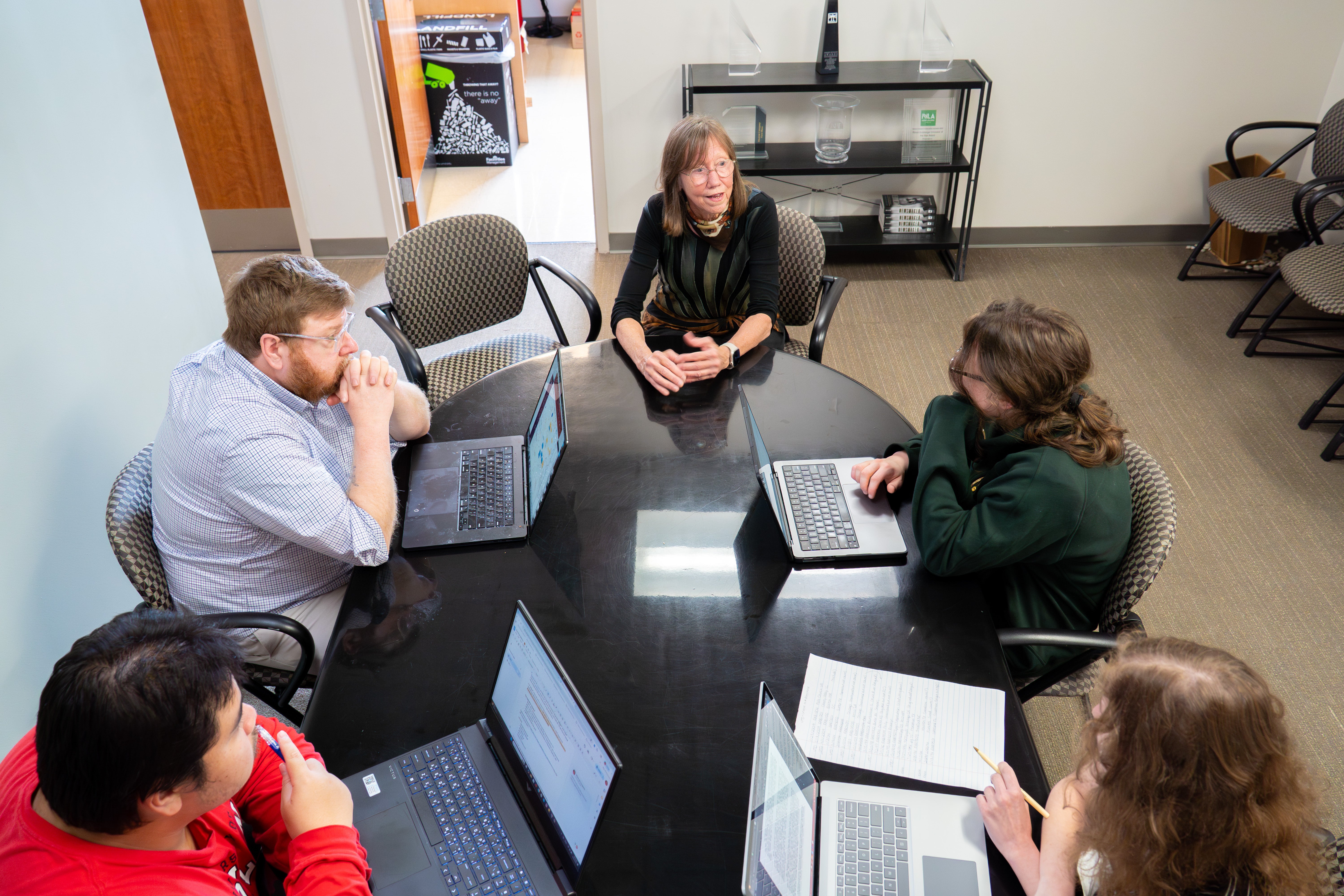 A group of five people sits around a conference table in a newsroom, working on laptops and reviewing documents during a meeting at the University of Maryland’s Howard Center for Investigative Journalism.