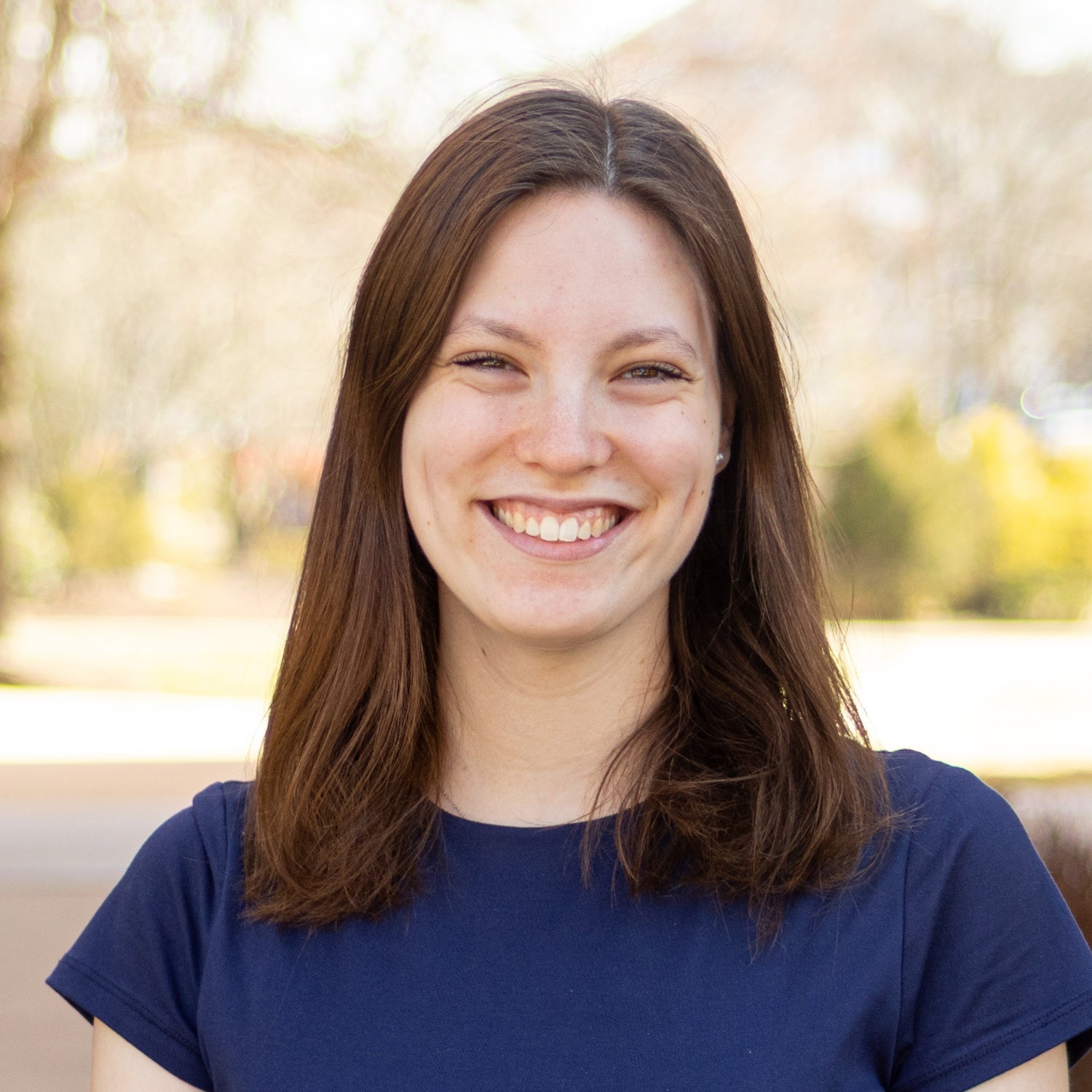 Portrait of Clare Roth outdoors on a sunny day, wearing a navy short-sleeve top, with shoulder-length brown hair and a softly blurred background of trees and campus buildings.