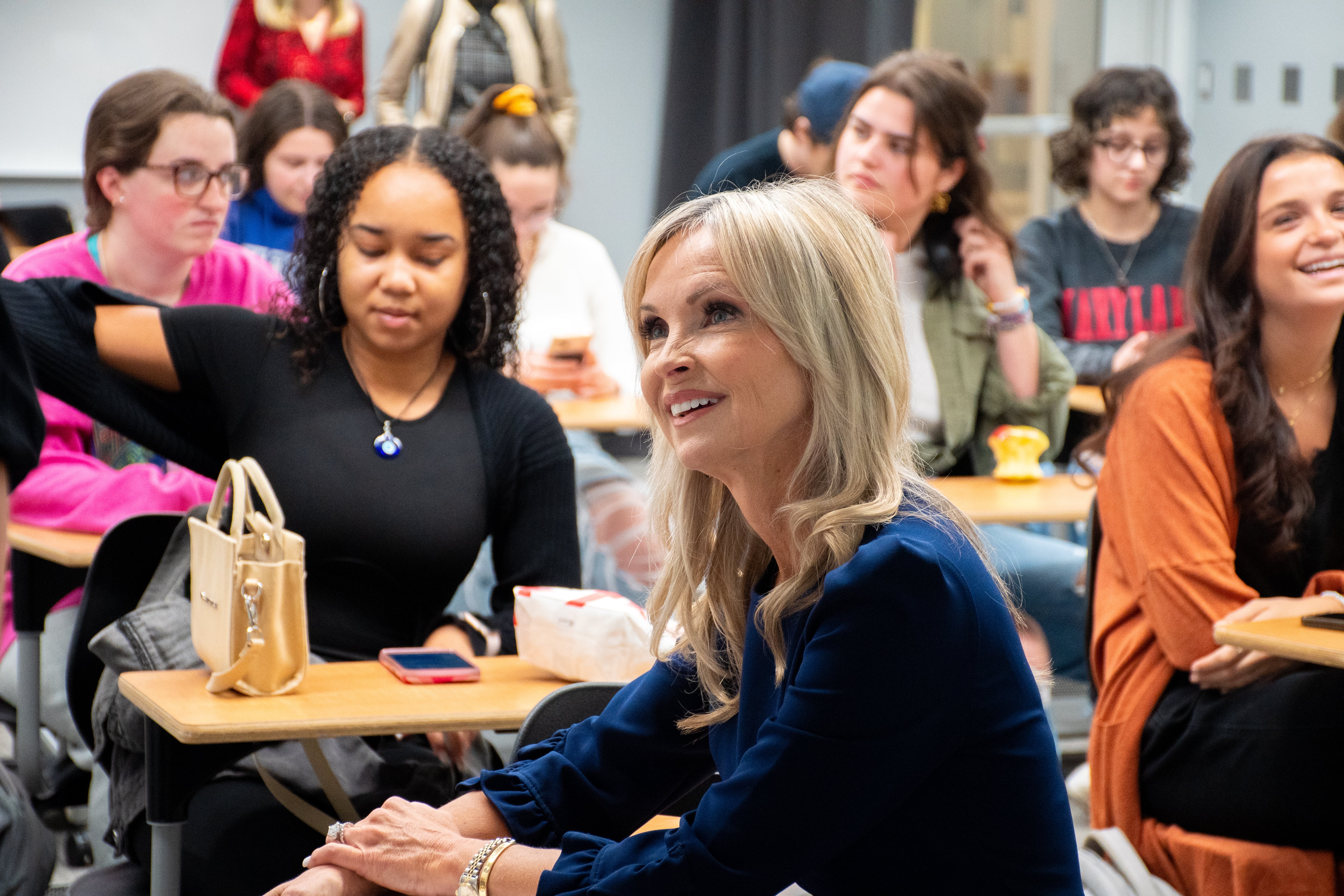 Jennifer Gilbert St. John sits at a classroom desk on the University of Maryland campus, smiling and engaged in conversation with students seated around her.