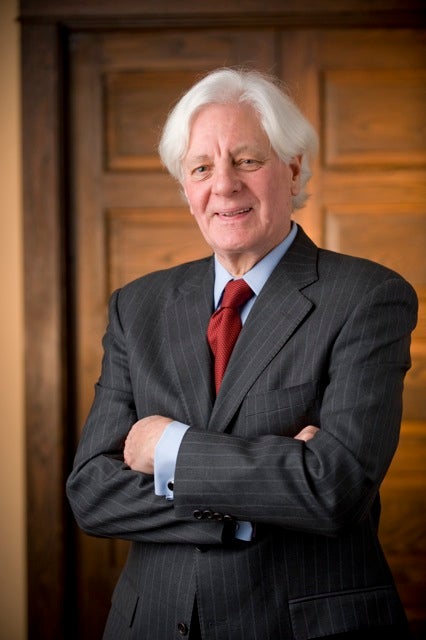 Portrait of Kevin Klose wearing a dark pinstripe suit and red tie, standing with arms crossed in front of a wood-paneled door.