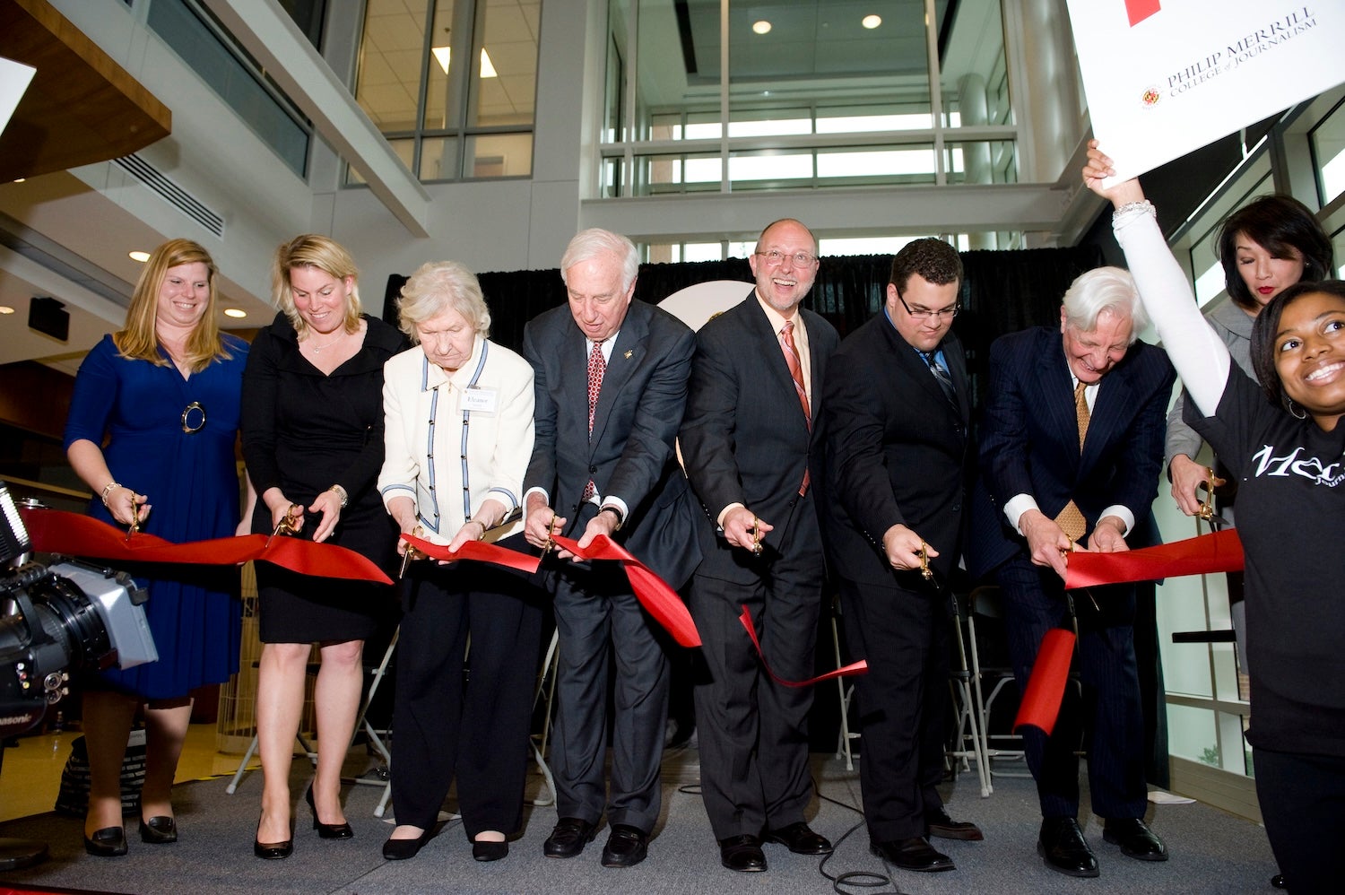 Group of University of Maryland and Merrill College officials participate in a ribbon-cutting ceremony for the dedication of Knight Hall, holding oversized scissors and cutting a long red ribbon inside the building’s glass-walled atrium.