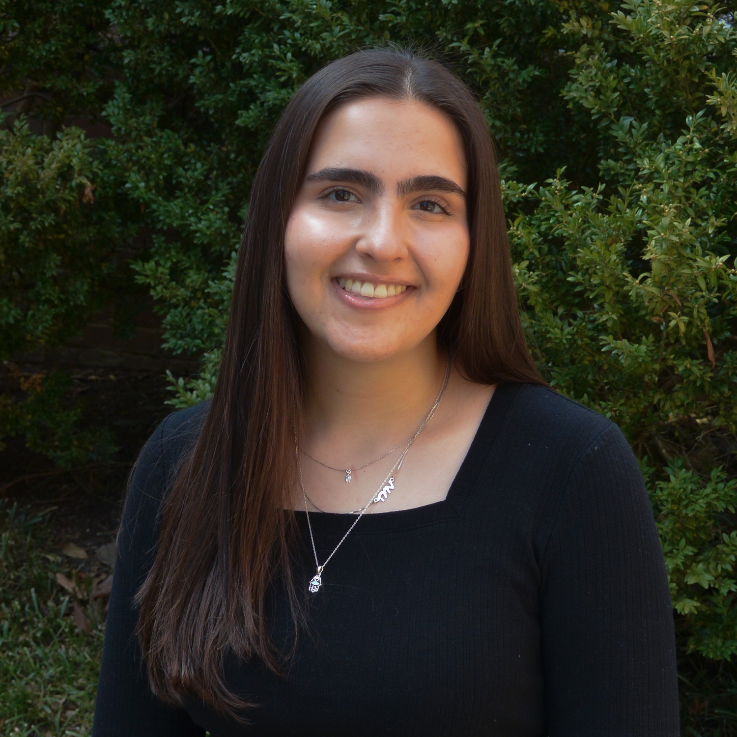 Portrait of Mayah Nachman standing outdoors in front of dense green shrubs, wearing a black long-sleeve top and layered silver necklaces, with long straight dark hair over one shoulder.