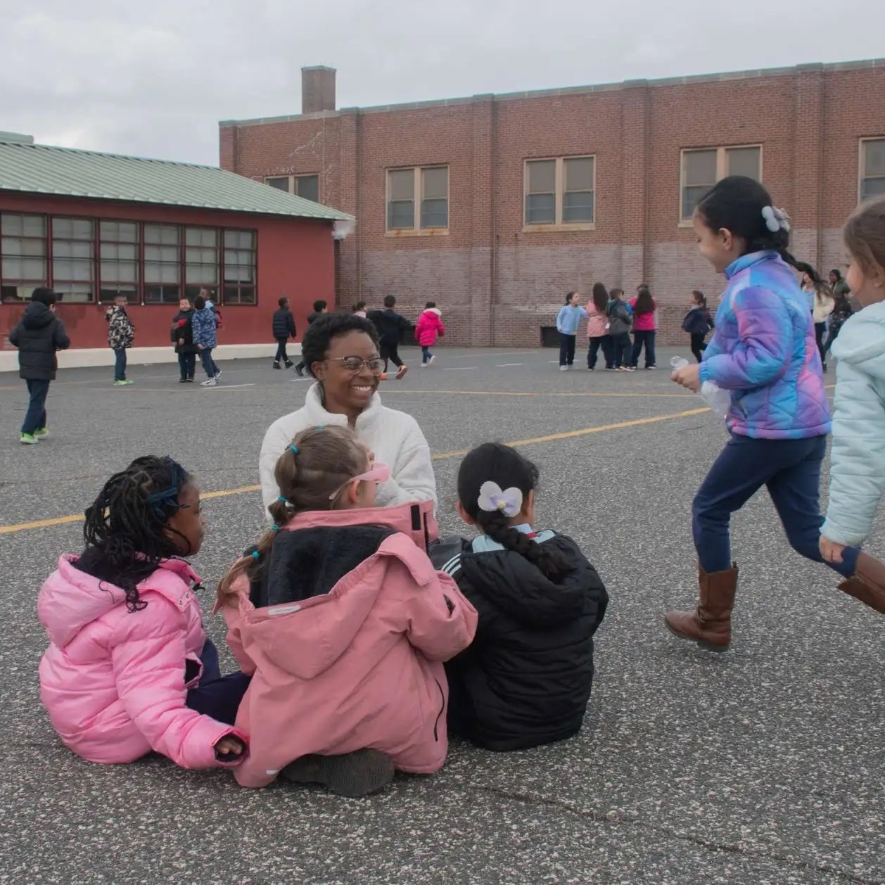 Children gather and play on an outdoor schoolyard, with several children seated in the foreground and others walking in the background near a brick school building. An adult stands among the children.