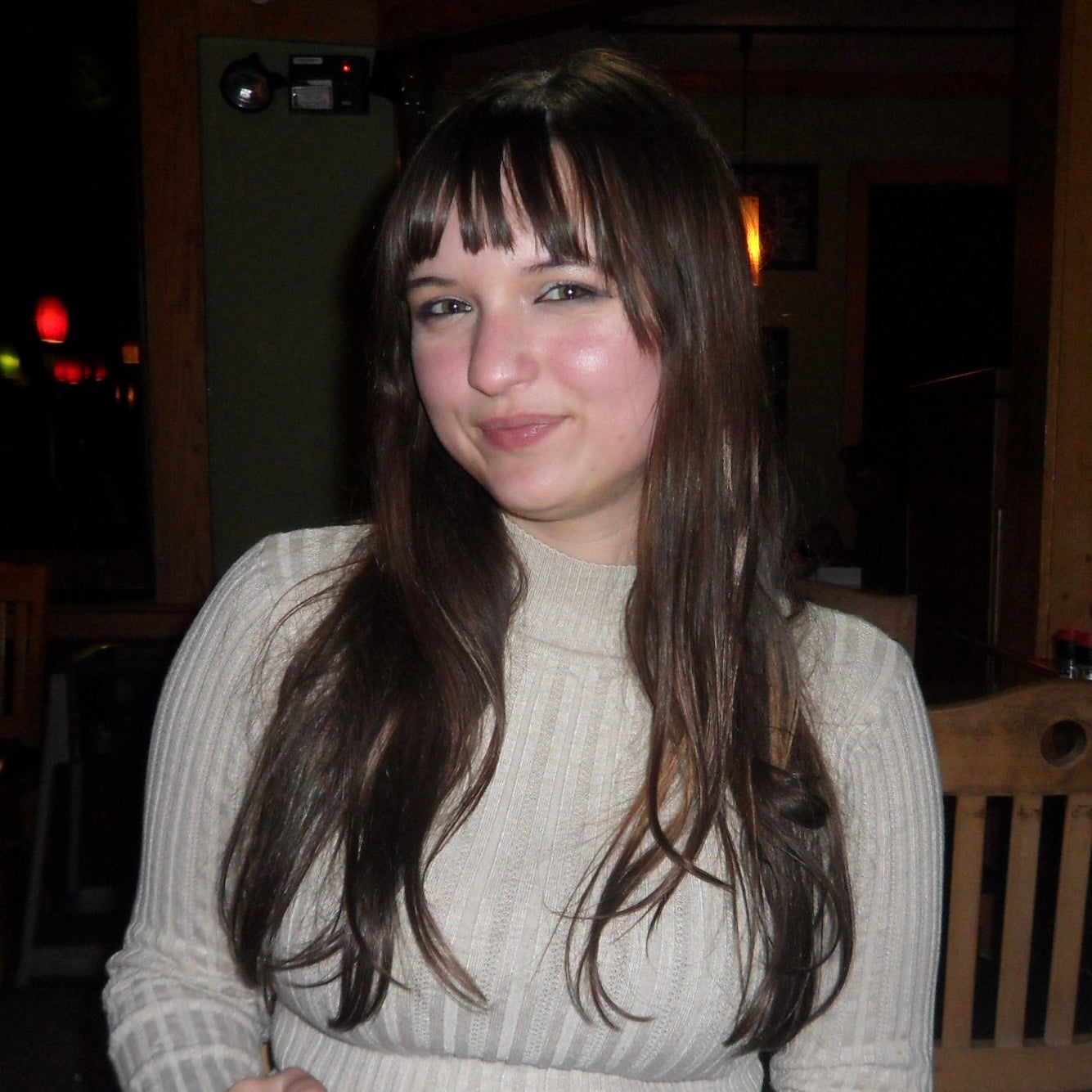 Portrait of Vada Mercer indoors in a dimly lit room, wearing a light gray ribbed sweater, with long dark hair over her shoulders and wooden chairs and warm lights in the background.