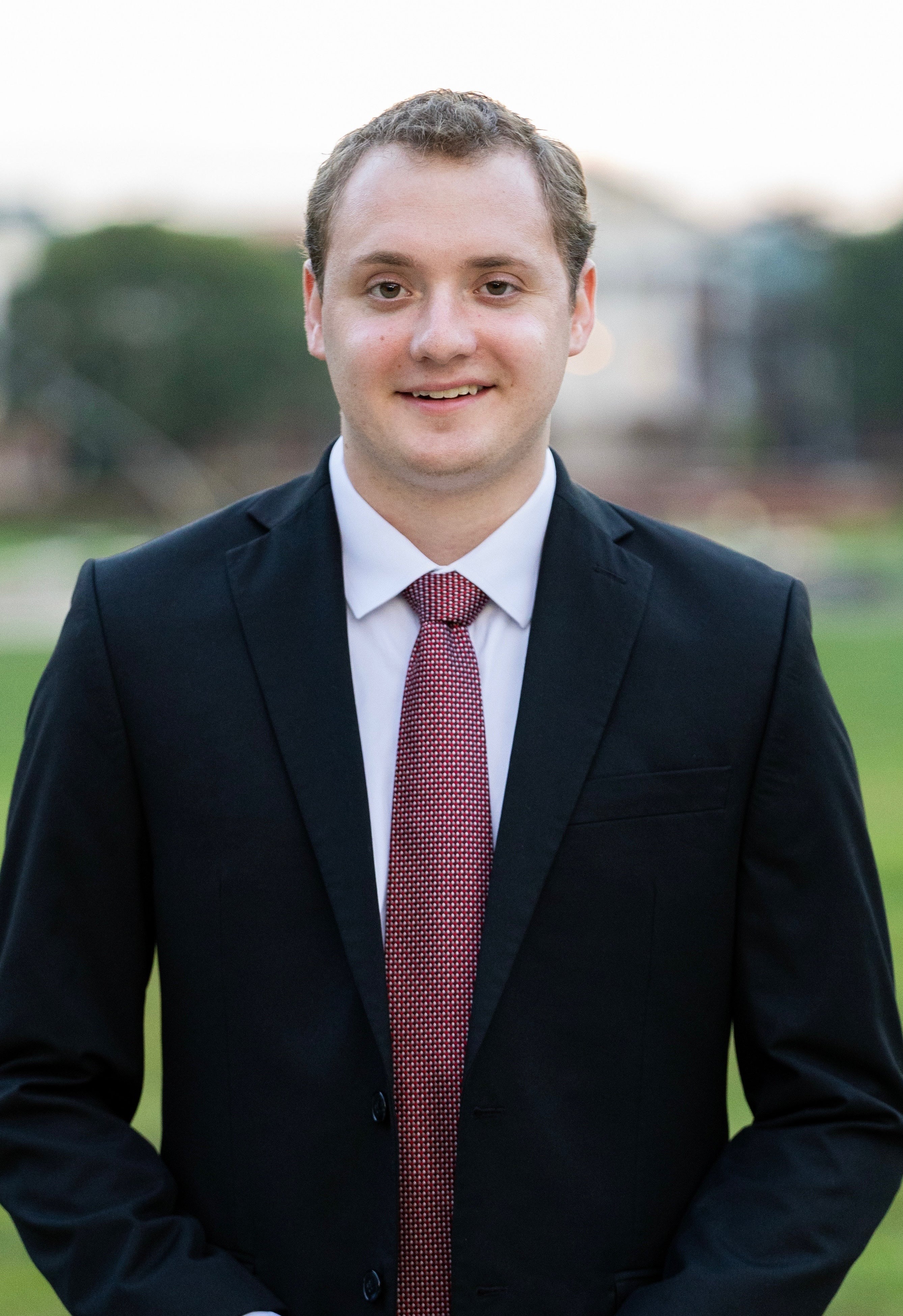 Portrait of Eddy Calkins smiling at the camera, wearing a black suit jacket, white dress shirt, and red patterned tie, standing outdoors with a softly blurred background of greenery and buildings.