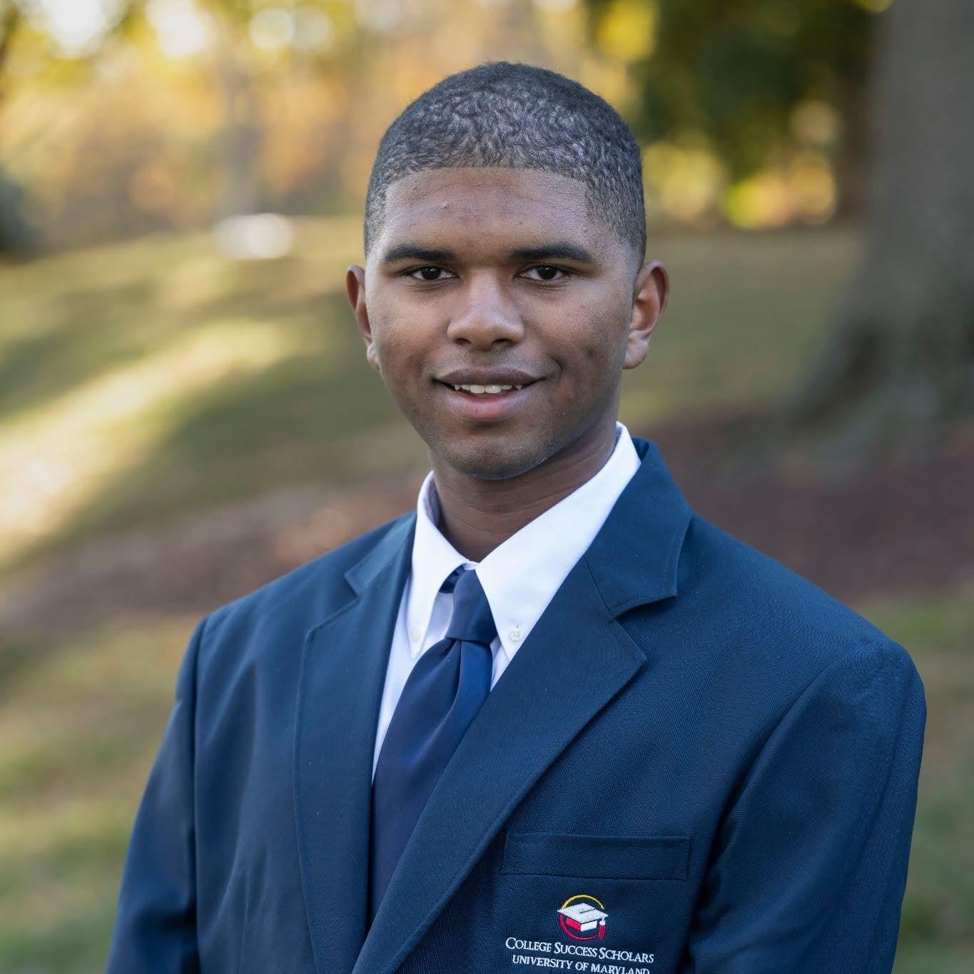 Portrait of Danny Chung-A-Fung standing outdoors in a park-like setting, wearing a navy suit jacket with a “College Success Scholars, University of Maryland” emblem, a white shirt, and a navy tie; trees and grass are softly blurred in the background.