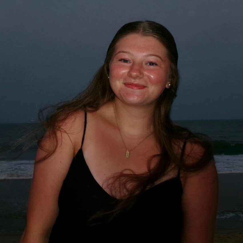 Portrait of Katherine Schutzman at the beach at dusk, wearing a black spaghetti-strap top and a small pendant necklace, with ocean waves and a darkening sky in the background.