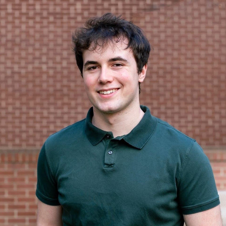 Portrait of Nolan Rogalski standing outdoors in front of a brick wall, wearing a dark green polo shirt, with the background softly blurred.