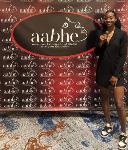 A young woman in a black blazer and shorts stands in front of an AABHE (American Association of Blacks in Higher Education) backdrop, smiling and holding a microphone. She wears black-and-white sneakers and a necklace. The backdrop features the AABHE logo and repeating branding, with a patterned carpet beneath her.