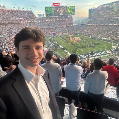 Harrison Rich smiling in a blazer and white collared shirt, taking a selfie from the press box overlooking a packed football stadium during the Super Bowl at Levi's Stadium.