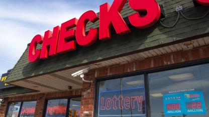 A convenience store exterior with a large red “CHECKS” sign above windows displaying lottery advertisements, including Powerball and Mega Millions jackpots.