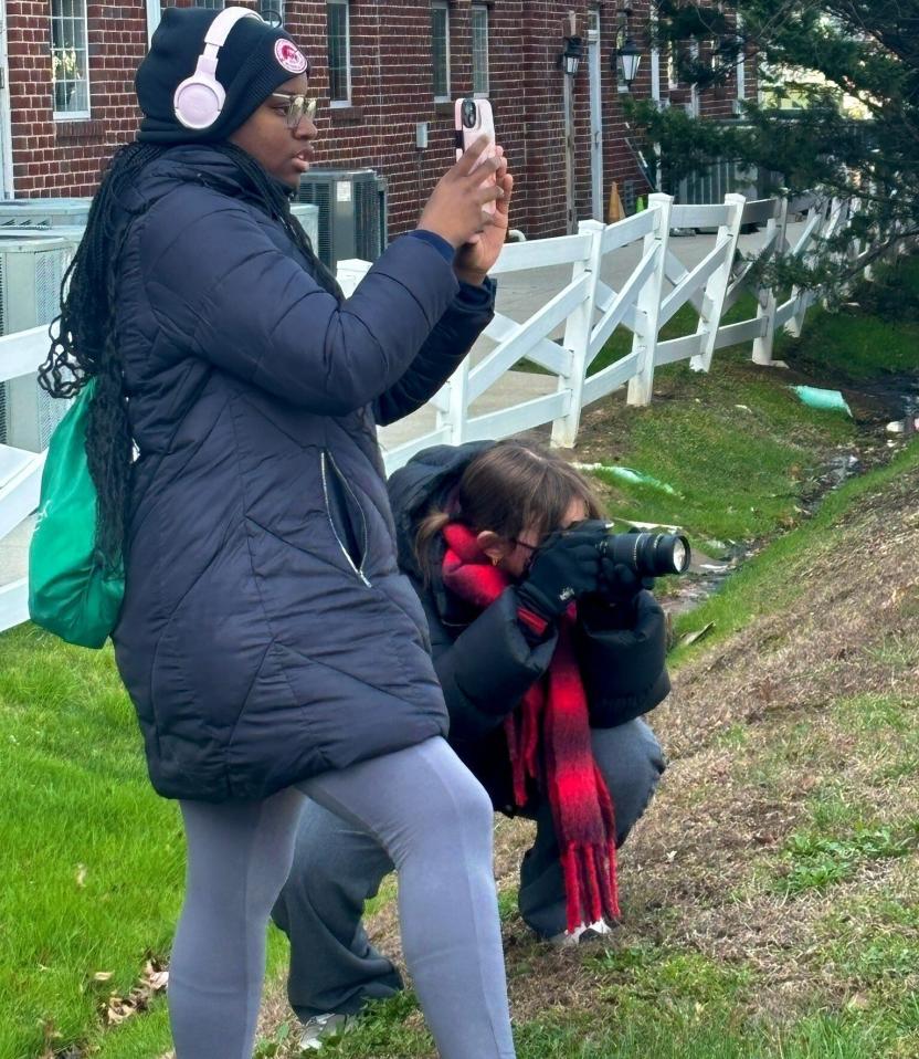 Two students stand on a grassy slope outdoors near a white fence and brick building, one taking a photo with a phone while the other crouches and photographs with a camera.