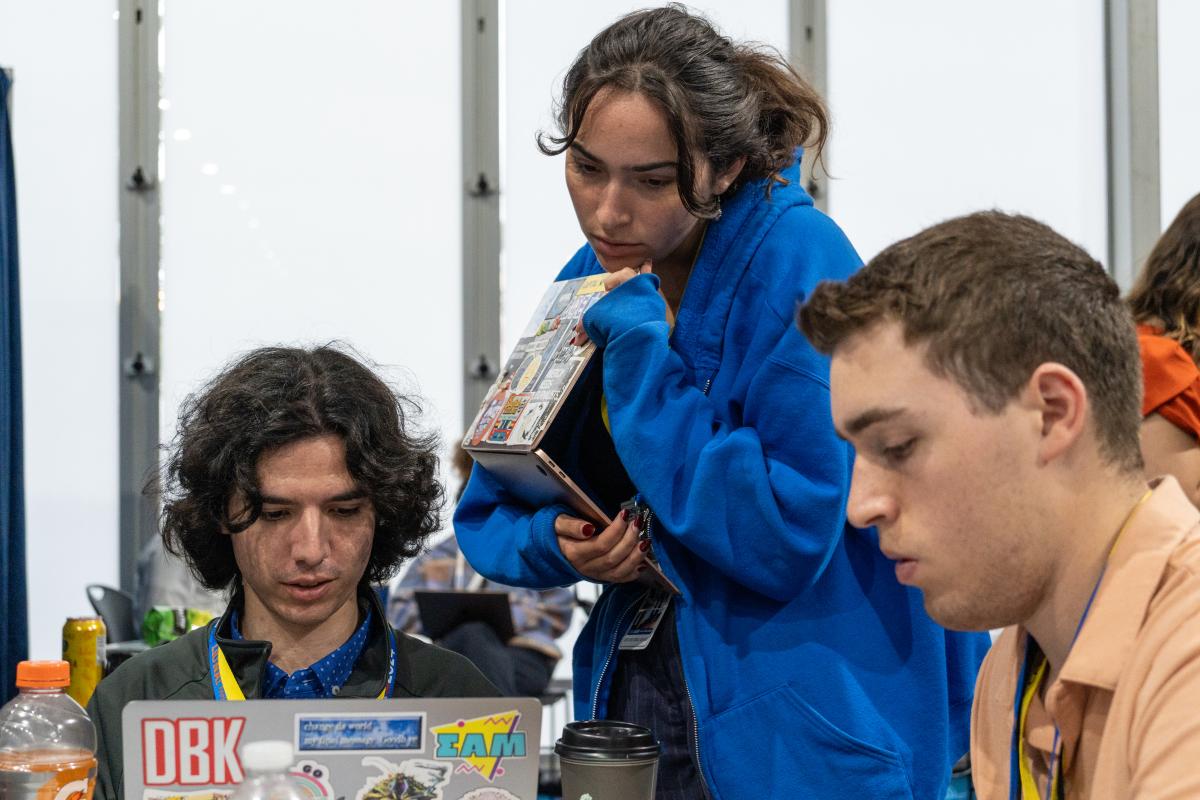 Three students collaborating on stories in front of computers in a newsroom setting.