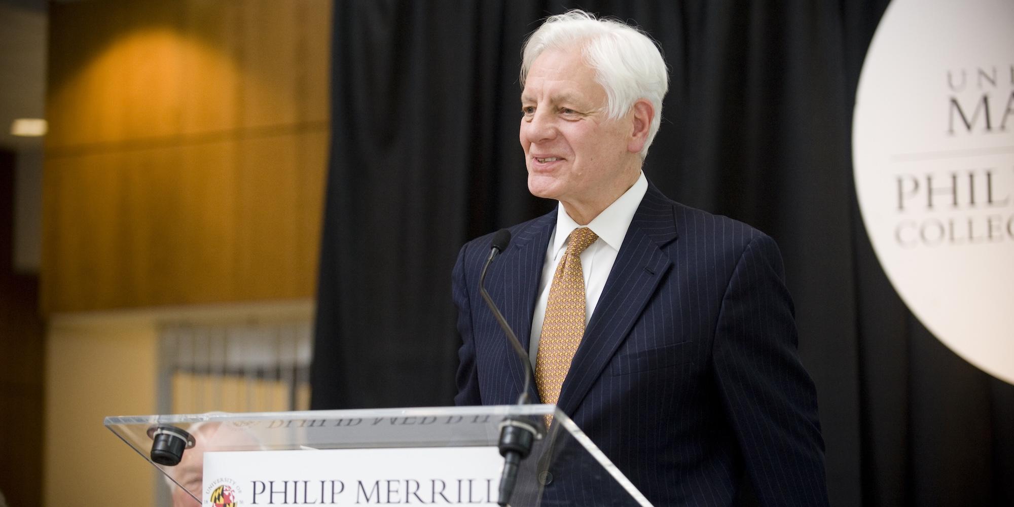 Kevin Klose stands at a podium labeled “Philip Merrill,” wearing a dark pinstripe suit and gold tie, speaking in front of black curtains and a University of Maryland Philip Merrill College of Journalism sign.