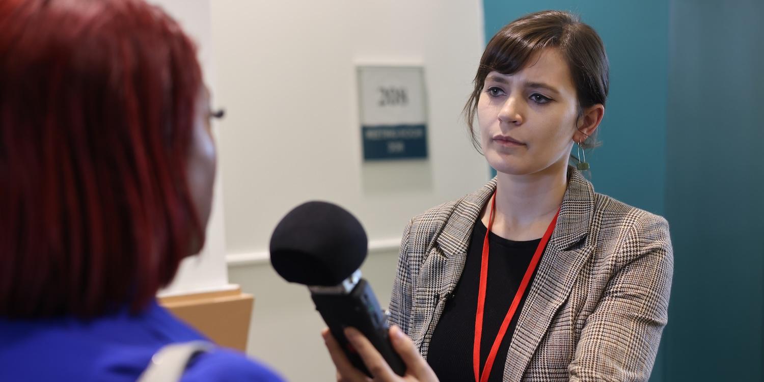 A reporter wearing a plaid blazer and red lanyard conducts an interview at a conference, holding a handheld microphone toward an attendee with bright red hair in a hallway outside meeting rooms.