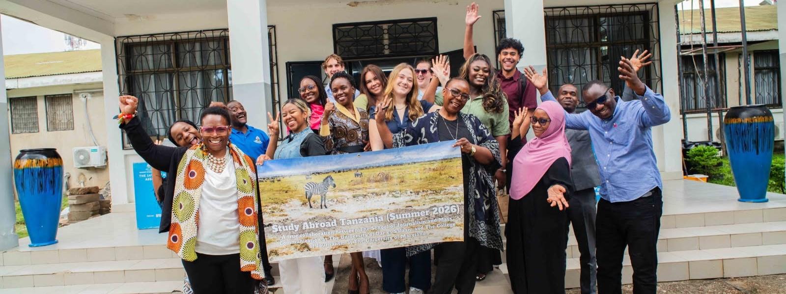 A group of people pose outside a building, smiling, waving, and holding a banner featuring zebras and the text “Study Abroad Tanzania (Summer 2026).” The banner describes a collaboration involving the University of Maryland’s Philip Merrill College of Journalism. The group includes people of diverse ages and backgrounds, with large blue decorative planters on either side of the entrance.