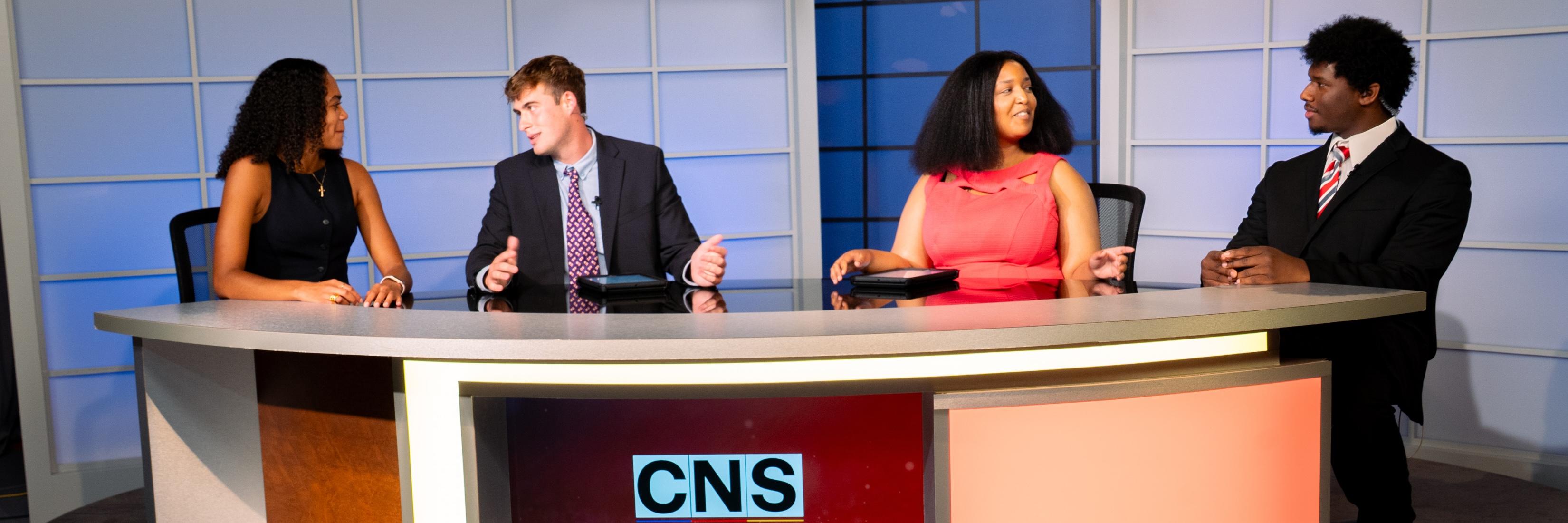Four student news anchors sit at a curved broadcast desk with the CNS logo in a television studio.