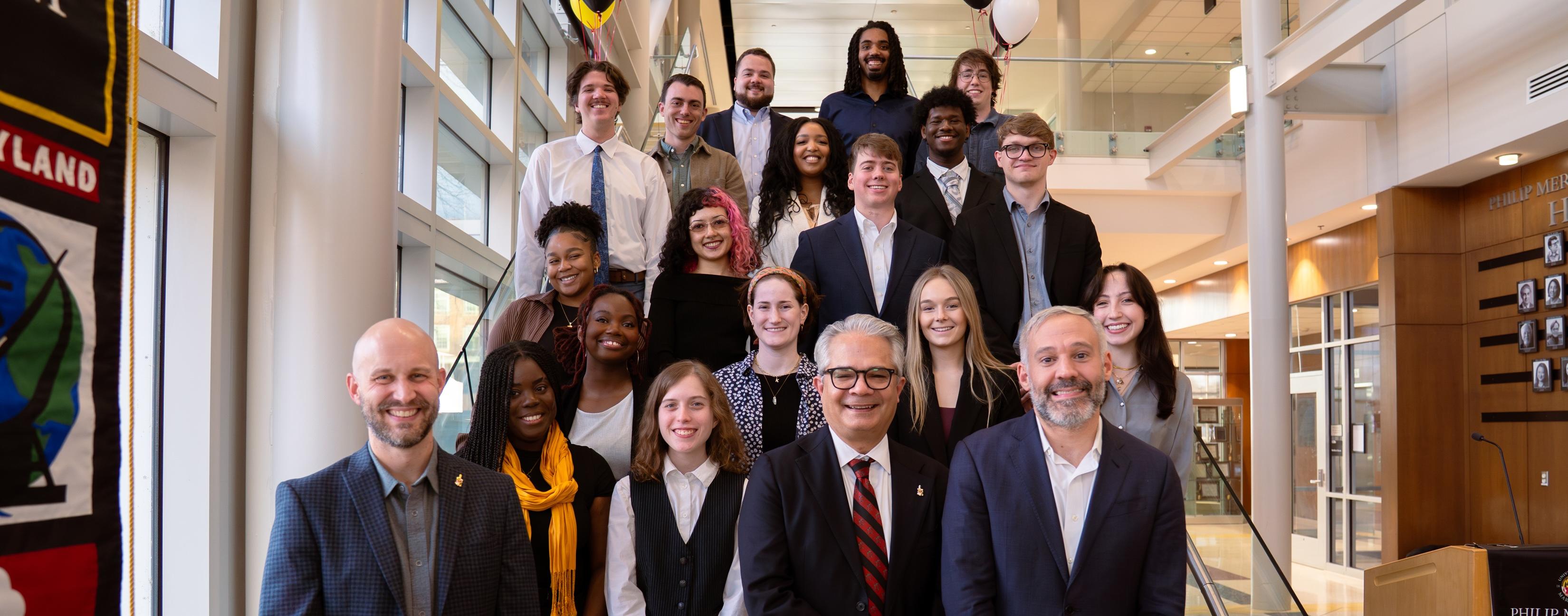 A group of 17 people pose for a group photo inside the Philip Merrill College of Journalism. Three people stand in the front, with students and faculty arranged on the steps behind them. Red, yellow, white and black balloons decorate the space, and a podium and college displays are visible in the background.