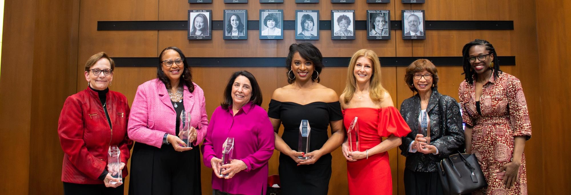 Seven elegantly dressed women smile and converse while holding crystal awards in front of a wood-paneled wall with framed portraits.