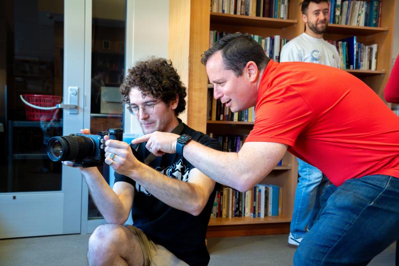 An instructor in a red shirt points out settings on a video camera screen for a student in a library or classroom.